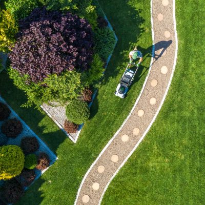 Large Beautiful Residential Backyard Garden Grass Mowing Aerial View. Caucasian Gardener Pushing Electric Cordless Grass Mower and Trimming the Lawn.