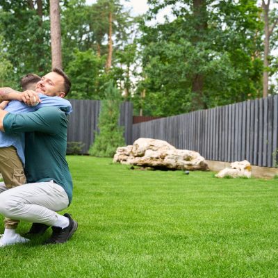 Smiling father hugs his son on the lawn of the house, the boy has a baseball glove